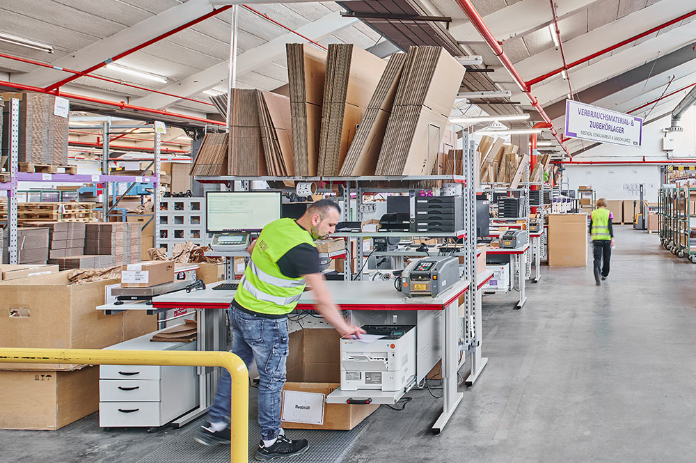 Packarbeitsplatz bei Müller – Die lila Logistik: ergonomischer Packtisch mit integriertem Zubehör, Kartons und Verpackungsmaterialien im Einsatz