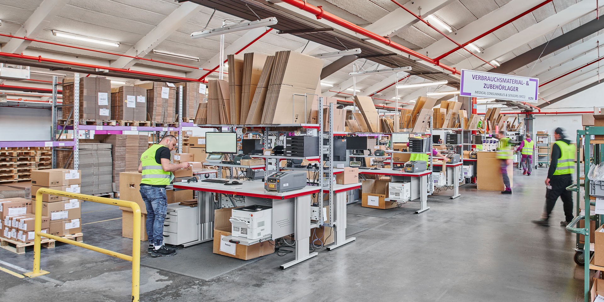 Moderner Logistik-Arbeitsbereich mit mehreren Packtischen und Lagerregalen im Hintergrund; helle, gut organisierte Halle mit Industriebeleuchtung