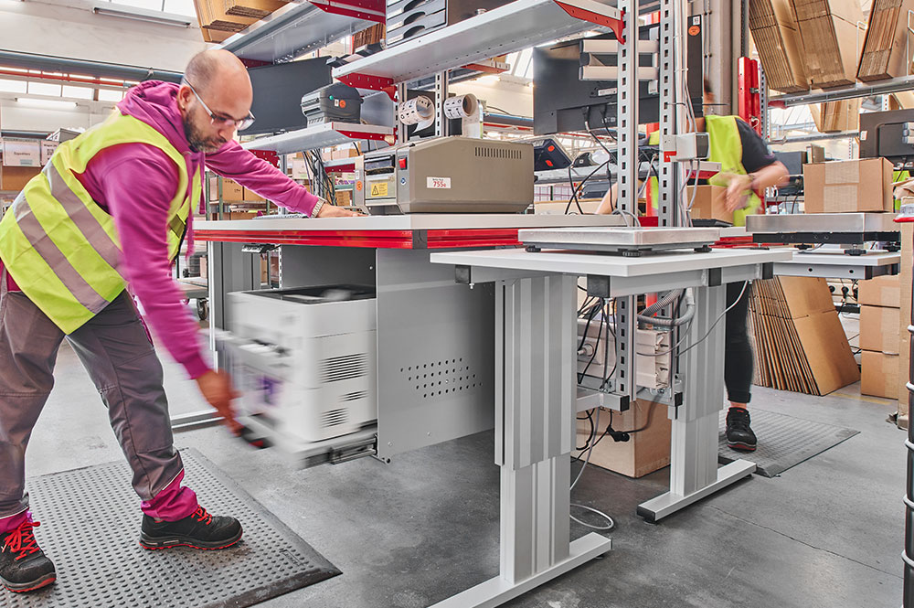 Mitarbeiter zieht den Druckerauszug eines Packarbeitsplatzes heraus; im Hintergrund sind Regale und technische Ausstattung zu sehen