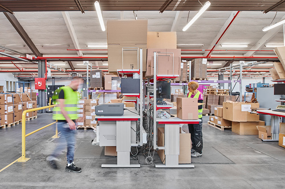 Packarbeitsplatz im Einsatz bei Müller – Die lila Logistik: Mehrere Mitarbeiter arbeiten an ergonomischen Packtischen