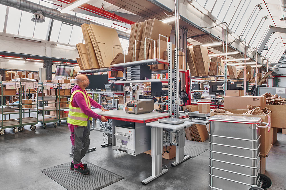 Detailansicht eines Packtischs bei Müller – Die lila Logistik: Strukturierter Arbeitsplatz mit Ablagefächern, Verpackungsmaterialien und integriertem Bildschirm